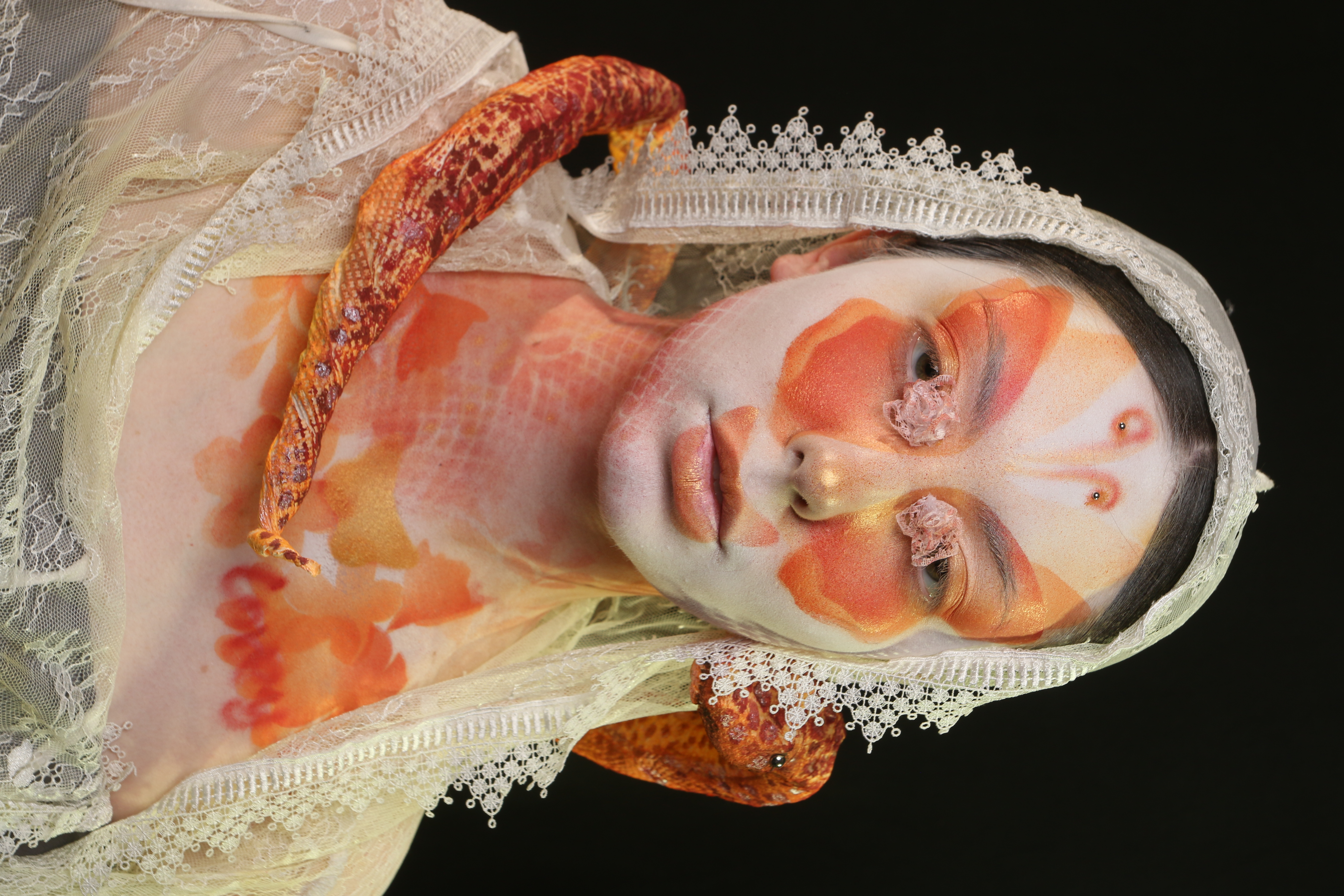 Side portrait of Lepidophis featuring orange butterfly-inspired makeup, white lace, and a serpent form surrounding the model.