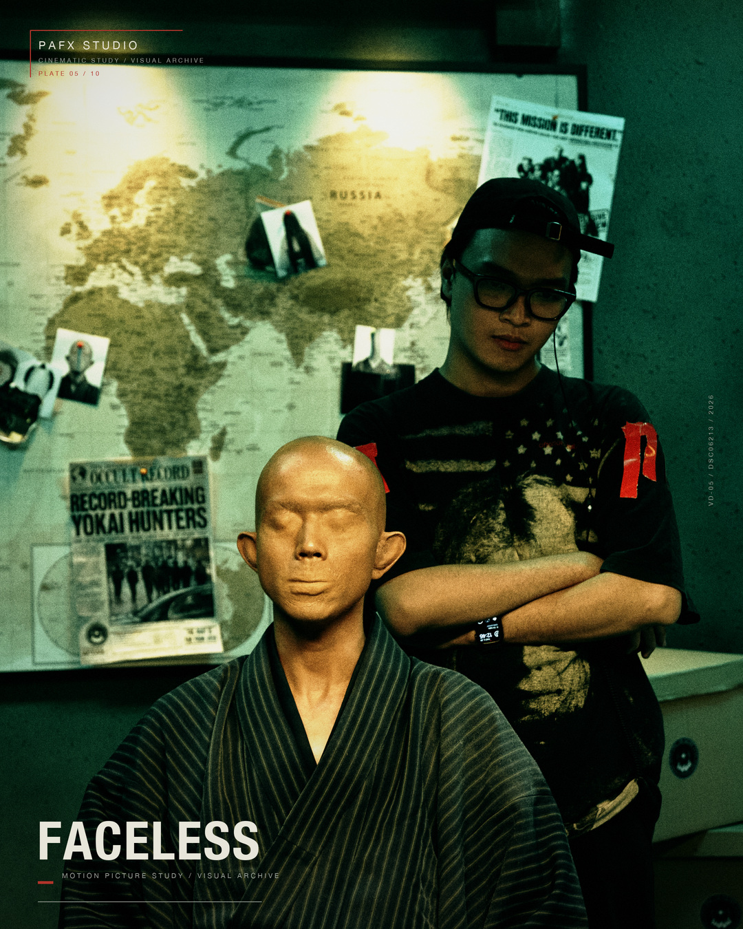 The FACELESS character seated against a map-covered set while a crew member works behind the performer.